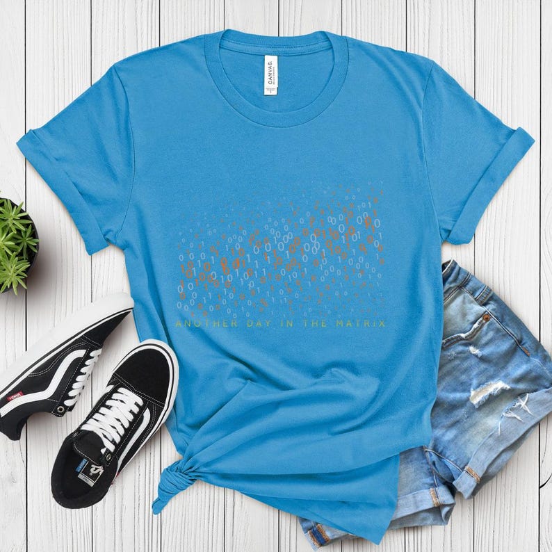 May include: A light blue t-shirt with a graphic of binary code and the text "ANOTHER DAY IN THE MATRIX" in green. The shirt is styled with black and white sneakers and denim shorts on a white wooden surface.
