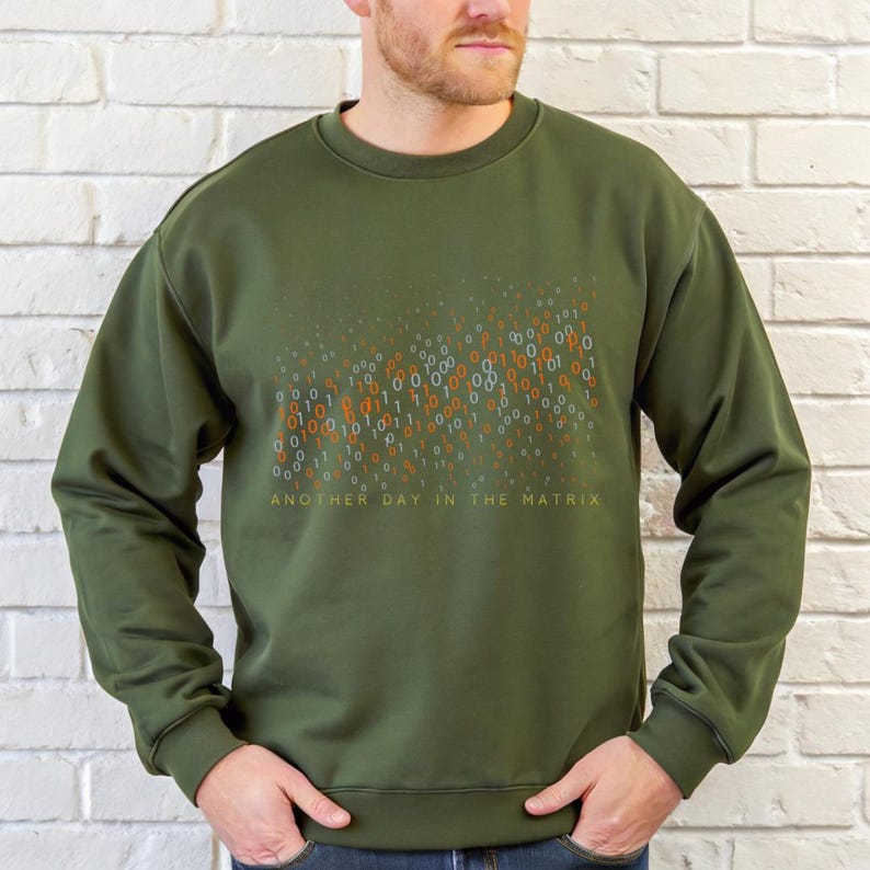 May include: Olive green sweatshirt with a binary code design in orange and teal. The text "ANOTHER DAY IN THE MATRIX" is printed below the graphic. The sweatshirt is a long-sleeved crew neck.