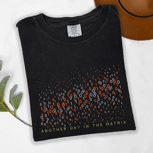 May include: Black t-shirt with a binary code design in orange, white, and blue. The text "ANOTHER DAY IN THE MATRIX" is printed in yellow at the bottom. The shirt has a crew neck and a Comfort Colors tag.