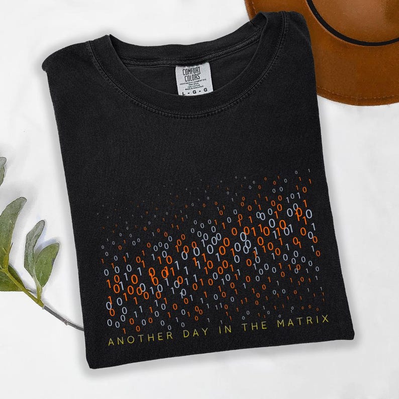 May include: Black t-shirt with a binary code design in orange, white, and blue. The text "ANOTHER DAY IN THE MATRIX" is printed in yellow at the bottom. The shirt has a crew neck and a Comfort Colors tag.