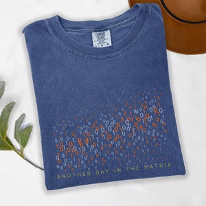 May include: A blue t-shirt with a binary code design in orange and white. The text "ANOTHER DAY IN THE MATRIX" is printed at the bottom in yellow. The shirt is made of a soft material and has a crew neck.