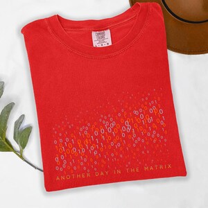 May include: A red t-shirt with the text "ANOTHER DAY IN THE MATRIX" in yellow. The shirt features a binary code design in white, orange, and yellow. The shirt is a Comfort Colors brand.