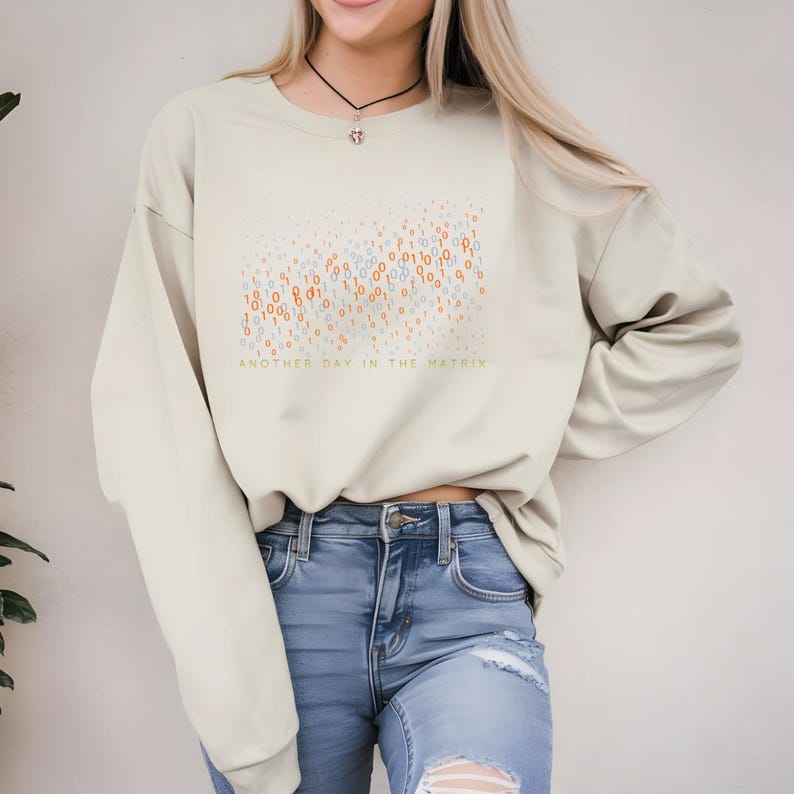May include: Beige sweatshirt with the text "ANOTHER DAY IN THE MATRIX" in green. The design features a pattern of orange, blue, and white binary code. The sweatshirt is paired with blue jeans.