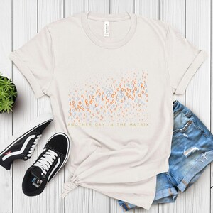May include: Off-white t-shirt with a binary code design in orange and blue, and the text "ANOTHER DAY IN THE MATRIX" in green. The shirt is styled with black and white sneakers and denim shorts.