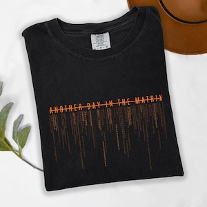 May include: Black t-shirt with the text "ANOTHER DAY IN THE MATRIX" in orange, along with a digital rain design. The shirt is folded and has a Comfort Colors tag.
