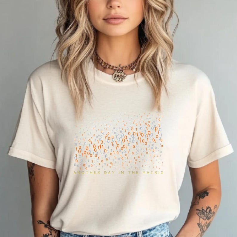 May include: Cream-colored t-shirt featuring a binary code design in orange and blue hues. The text "ANOTHER DAY IN THE MATRIX" is printed below the graphic. The shirt has short sleeves and a crew neck.
