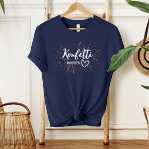 May include: Navy blue t-shirt with the text "Konfetti im Herzen" in white, surrounded by colorful confetti. The shirt is knotted at the waist and hangs on a wooden hanger.