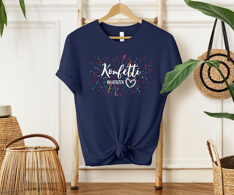 May include: Navy blue t-shirt with the text "Konfetti im Herzen" in white, surrounded by colorful confetti. The shirt is knotted at the waist and hangs on a wooden hanger.