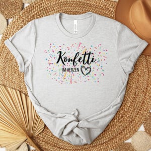 May include: A light gray t-shirt with the text "Konfetti im Herzen" in black script, surrounded by colorful confetti. A black heart is below the text. The shirt is made of a soft material.