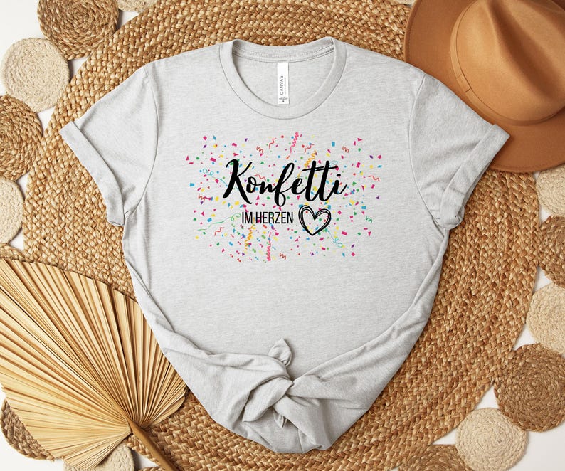 May include: A light gray t-shirt with the text "Konfetti im Herzen" in black script, surrounded by colorful confetti. A black heart is below the text. The shirt is made of a soft material.