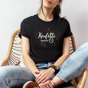 May include: Black t-shirt with the text "Konfetti im Herzen" in white script, featuring a heart symbol. The design includes colorful confetti-like accents. The shirt is worn by a person sitting in a wicker chair.
