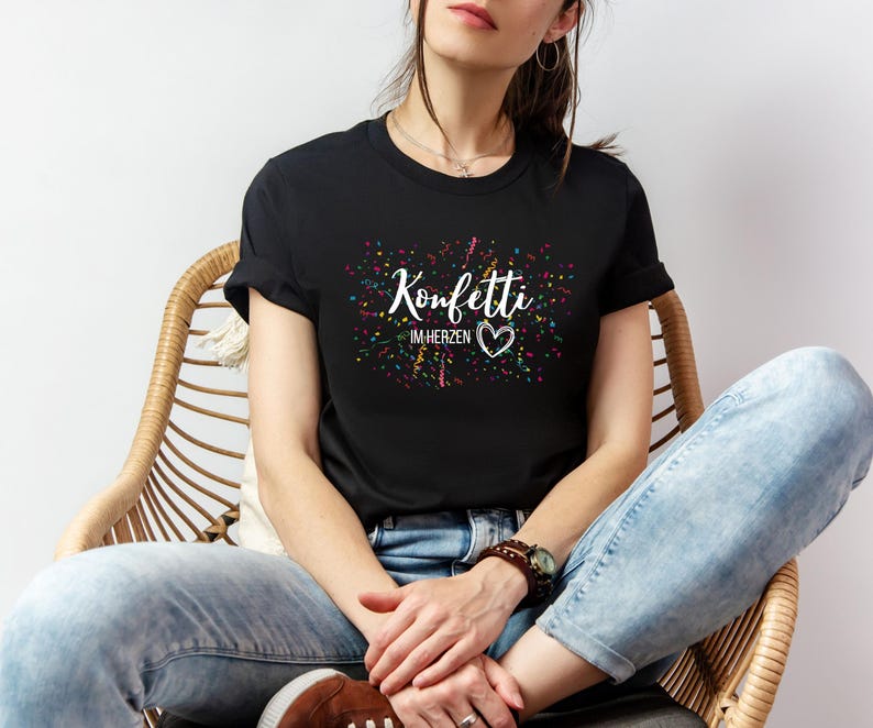 May include: Black t-shirt with the text "Konfetti im Herzen" in white script, featuring a heart symbol. The design includes colorful confetti-like accents. The shirt is worn by a person sitting in a wicker chair.