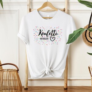 May include: White t-shirt with the text "Konfetti im Herzen" in black script, a heart, and colorful confetti. The shirt is hanging on a wooden rack. A woven basket and a straw bag are in the background.