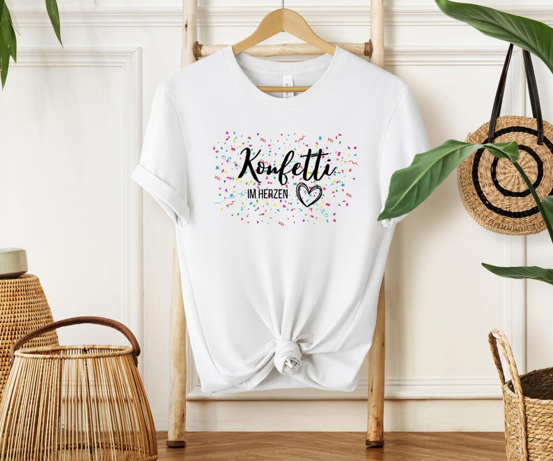 May include: White t-shirt with the text "Konfetti im Herzen" in black script, a heart, and colorful confetti. The shirt is hanging on a wooden rack. A woven basket and a straw bag are in the background.