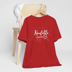 May include: A red t-shirt with the words "Konfetti im Herzen" in white script, surrounded by colorful confetti. A white heart is below the text. A white baseball cap rests on a wooden chair.