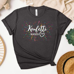 May include: Dark gray t-shirt with the text "Konfetti im Herzen" in white script, a heart, and colorful confetti. The shirt is made of a soft material and has rolled-up sleeves. A casual, everyday tee.