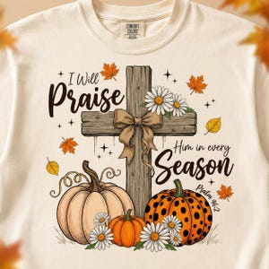 May include: A cream-colored crewneck sweatshirt featuring a graphic of a wooden cross with a tan bow, surrounded by pumpkins, white daisies, and autumn leaves. The text reads I Will Praise Him in every Season and Psalm 46:2 in black script.