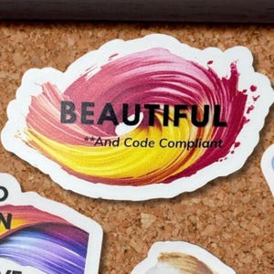 May include: A white sticker with the word "BEAUTIFUL" in black text, set against a swirl of red and yellow paint strokes. The text "And Code Compliant" is below the main word.