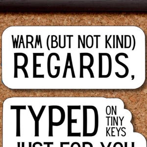 May include: Two white rectangular stickers with black text on a corkboard background. The top sticker reads "WARM (BUT NOT KIND) REGARDS,". The bottom sticker says "TYPED ON TINY KEYS".