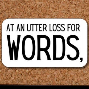 May include: A white rectangular sticker with rounded corners, featuring the phrase "AT AN UTTER LOSS FOR WORDS," in black, bold font. The sticker is displayed against a corkboard background.