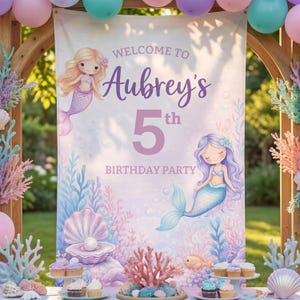 May include: A mermaid-themed birthday party backdrop with the text "Welcome to Aubrey's 5th Birthday Party." The backdrop features illustrations of mermaids, seashells, coral, and cupcakes. Balloons in pastel colors decorate the top.