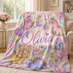May include: A soft plush blanket featuring a watercolor print of various pastel-colored dinosaurs, flowers, and butterflies. The name Olivia is written in large, cursive purple script across the center of the blanket, surrounded by illustrations of dinosaurs.