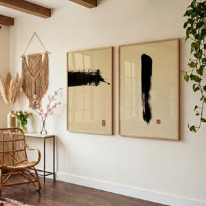 May include: Two framed abstract art prints. One features a black brushstroke, the other a vertical black stroke. Both are on a cream background with a small red stamp. The frames are light wood.