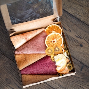 Pode incluir: Uma caixa de cartão com uma janela transparente, cheia de triângulos de couro de fruta coloridos e fatias de fruta seca. O couro de fruta é em tons de rosa, vermelho e castanho. Fatias de laranja e kiwi secas também são visíveis.