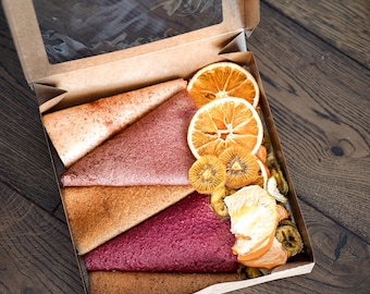 Op de afbeelding: Een kartonnen doos met een doorzichtig venster, gevuld met kleurrijke fruitlederdriehoeken en gedroogde fruitschijfjes. Het fruitleer is in tinten roze, rood en bruin. Gedroogde sinaasappel- en kiwischijfjes zijn ook zichtbaar.