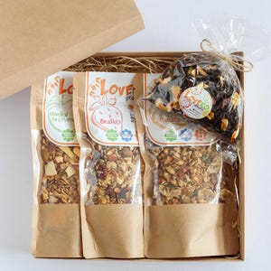 Op de afbeelding: Een geschenkdoos met drie zakken granola en een zak gedroogd fruit. De granolazakken zijn voorzien van het label "Love" met verschillende fruitnamen. De zak met gedroogd fruit is vastgebonden met touw. De doos is bruin.