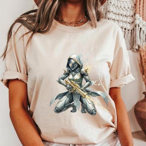 May include: Beige t-shirt featuring a watercolor illustration of a futuristic warrior in silver armor, kneeling and holding a golden weapon. The warrior has a hooded helmet and is set against a backdrop of yellow and white splashes.