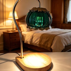 May include: A table lamp featuring a curved metallic arm and a scalloped, emerald green glass shade. The base is a light-toned, circular wooden platform that emits a warm, bright light from its surface, illuminating the area beneath the lamp.