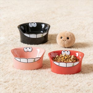 May include: Three ceramic pet bowls in black, pink, and red, each with a smiling face design. The red bowl is filled with pet food. A small, plush, round toy with a smiling face is also present.