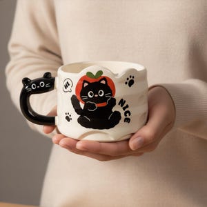 Puede incluir: Taza de cerámica blanca con asa negra en forma de cabeza de gato. La taza presenta un gato negro de dibujos animados con un sombrero de tomate rojo, huellas de patas y la palabra "NICE". El asa tiene el texto "0.0=".