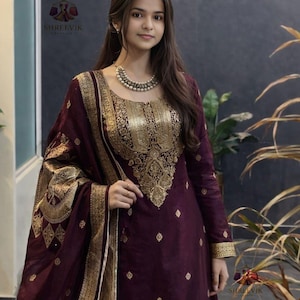 May include: A plum-colored salwar kameez ensemble with intricate gold embroidery on the neckline, sleeves, and dupatta. The outfit features a detailed gold pattern and is worn with a matching scarf. The image includes the logo "SHREEVIK".