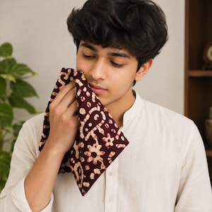 Op de afbeelding: Een vierkante stoffen zakdoek met een diep kastanjebruine achtergrond en een herhalend crème-kleurig bloemen- en paisley-patroon in batikstijl. De doek is gevouwen en wordt vastgehouden, wat het complexe, handgeverfde uiterlijk van het textielontwerp toont.
