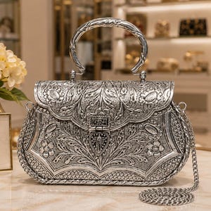 May include: A silver-toned metal handbag featuring an intricate embossed floral pattern across the front flap and body. The bag has a rounded top handle, a central clasp closure, and a detachable thin silver chain shoulder strap.