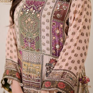 May include: A long-sleeved, light pink tunic with intricate patchwork design. The tunic features floral and geometric patterns in shades of purple, green, and yellow, with mirrored accents. The sleeves are adorned with floral borders and dangling embellishments.