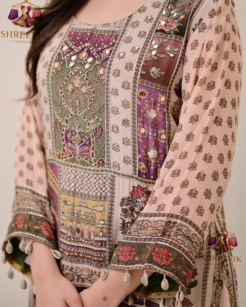 May include: A long-sleeved, light pink tunic with intricate patchwork design. The tunic features floral and geometric patterns in shades of purple, green, and yellow, with mirrored accents. The sleeves are adorned with floral borders and dangling embellishments.