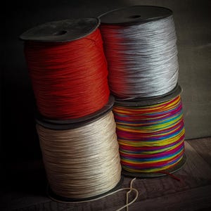 May include: A stack of four spools of cord. The top spool is red, the next is silver, followed by a beige spool, and the bottom spool is multi-colored. A length of beige cord is in the foreground. The spools are on a dark wooden surface.