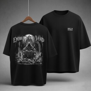 May include: Black t-shirts with a gothic design. One shirt features the text "EMBRACE THE VOID" with an illustration of a cloaked figure on a throne of skulls. The other shirt has the text "BUILT IN SILENCE" on the front.