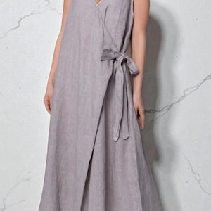 May include: A lavender-coloured wrap dress with a V-neck and tie closure. The sleeveless dress is made of a lightweight fabric, possibly linen. The dress is designed in a wrap style, with the fabric overlapping at the front and tying at the side.