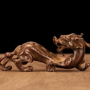 Op de afbeelding: Een bronzen, gestileerd drakenbeeldje. Het mythische wezen wordt afgebeeld in een dynamische pose, met een gebogen lichaam en een geheven hoofd. De sculptuur heeft ingewikkelde details, waaronder schubben en gelaatstrekken. De achtergrond is een houten oppervlak met een zwarte achtergrond.