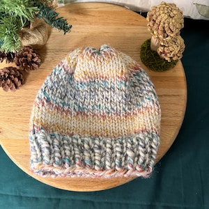 May include: A hand-knitted beanie hat with a ribbed brim. The hat features a mix of colors, including cream, blue, pink, and yellow. The hat is made of a thick, textured yarn, perfect for cold weather.