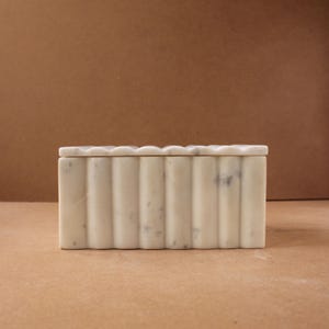 Puede incluir: Una caja rectangular de mármol blanquecino con tapa festoneada. La caja tiene crestas verticales y redondeadas a lo largo de los lados. El mármol tiene sutiles vetas grises. La caja es un artículo de almacenamiento decorativo.