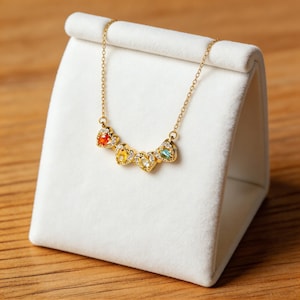 May include: A gold-colored necklace with five heart-shaped pendants, each with a different colored gemstone. The necklace is displayed on a white velvet stand. The gemstones are red, yellow, light green, and blue.