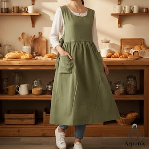 May include: An olive green apron with two large front pockets. The apron is worn over a white long-sleeved shirt and blue jeans. The apron has a gathered waist and ties at the sides. The model is wearing white sneakers.
