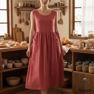 May include: A sleeveless, dusty rose-colored dress with a round neckline and two large front pockets. The dress has a gathered waist and tie details at the sides. The dress is made of a soft, flowing fabric.