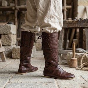 Könnte beinhalten: Ein Paar dunkelbraune, kniehohe Stiefel mit einer Schnürung, die sich um die Wade wickelt und oben gebunden wird. Die Stiefel haben eine glatte Oberfläche, eine abgerundete Zehenpartie und eine flache Sohle, getragen zu einer hellen, weiten Hose.