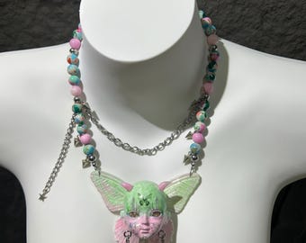 May include: A unique necklace featuring a pendant with a face and butterfly wings in shades of green and pink. The necklace has a beaded strand with pink, blue, and green beads, and a silver chain. The pendant has dangling earrings.